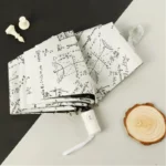 Parapluie beige pliant automatique motif formules mathématiques, anti-UV, posé sur une surface claire avec accessoires décoratifs.