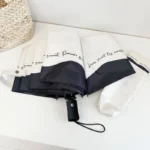 Parapluie blanc bord noir anti-UV déplié, design élégant avec étui, sur surface claire près d'un panier en osier.