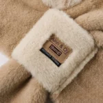 Capuche hiver beige en fausse fourrure avec écharpe intégrée, détail étiquette cuir 'Alinn & Qy Est. 1987' pour look chic et confort.