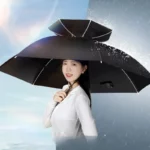 Femme souriante avec chapeau parapluie noir grande taille, protection pluie élégante, ciel bleu et gouttes d'eau en arrière-plan.