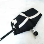 Parapluie de luxe plié noir et blanc avec motifs élégants, manche ergonomique noir et chaînette décorative sur fond neutre.