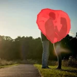 Couple sous un parapluie rouge cœur à froufrou, chemin nature au coucher de soleil, symbole d’amour et élégance romantique.