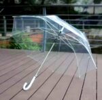 Parapluie transparent avec poignée blanche, design élégant et pratique, structure métallique, posé sur un sol en bois.