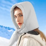 Femme avec manteau et capuche beige matelassée, neige et ciel bleu en fond, accessoire hiver élégant et chaud.