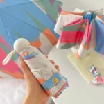 Mini parapluie manuel anti-UV pop art coloré, motif artistique rose, bleu, orange, plié sur une table avec un autre ouvert derrière.