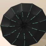 Parapluie anti retournement élégant, collection moderne, noir avec accents turquoise, ouvert sur une surface beige.