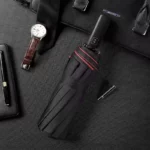 Parapluie rouge et noir luxe, pliable et automatique, élégamment posé avec une montre, un carnet en cuir et un stylo sur fond sombre.