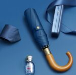 Parapluie homme pliant bleu avec poignée bois, cravate assortie, boîte de rangement et flacon de parfum sur fond bleu.
