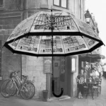 Parapluie cloche transparent Le Vieux Paris, décoré de motifs urbains, dans une rue animée avec cafés et passants en arrière-plan.