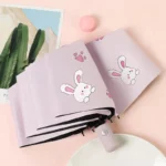 Parapluie pliant enfant rose lapin anti-UV, illustrations pattes, idéal pour protection mode, fond rose décor carte et macaron.