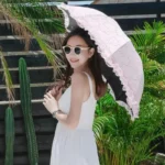 Femme en robe blanche avec un parapluie pliant rose à dentelle pour mariage, posant en extérieur près de plantes tropicales.
