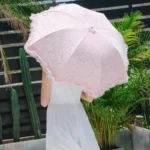 Parapluie rose en dentelle tenu par une personne en robe blanche, avec plantes tropicales, alliant élégance et protection solaire.