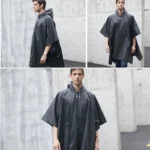 Homme portant un poncho de pluie noir taille unique, debout dans un cadre urbain moderne avec murs en béton.