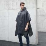 Homme avec poncho de pluie noir, taille unique, style moderne, idéal pour la pluie, en décor urbain avec murs en béton.