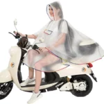 Femme en poncho transparent blanc avec capuche, poche intégrée, roulant en scooter beige sous la pluie, look pratique et stylé.