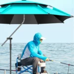 Pêcheur au bord de l'eau sous un parapluie octogonal turquoise et noir, équipé pour la pêche avec veste à capuche et lunettes.