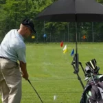 Parapluie golf clip noir protégeant un golfeur et son équipement sur un parcours avec drapeaux colorés en arrière-plan.