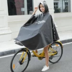 Femme souriante portant un poncho pluie large gris sur un vélo jaune, parfait pour rester au sec à vélo ou scooter.