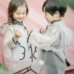 Deux enfants joyeux portant des ponchos transparents à boutons, jouant dans un cadre coloré et convivial.